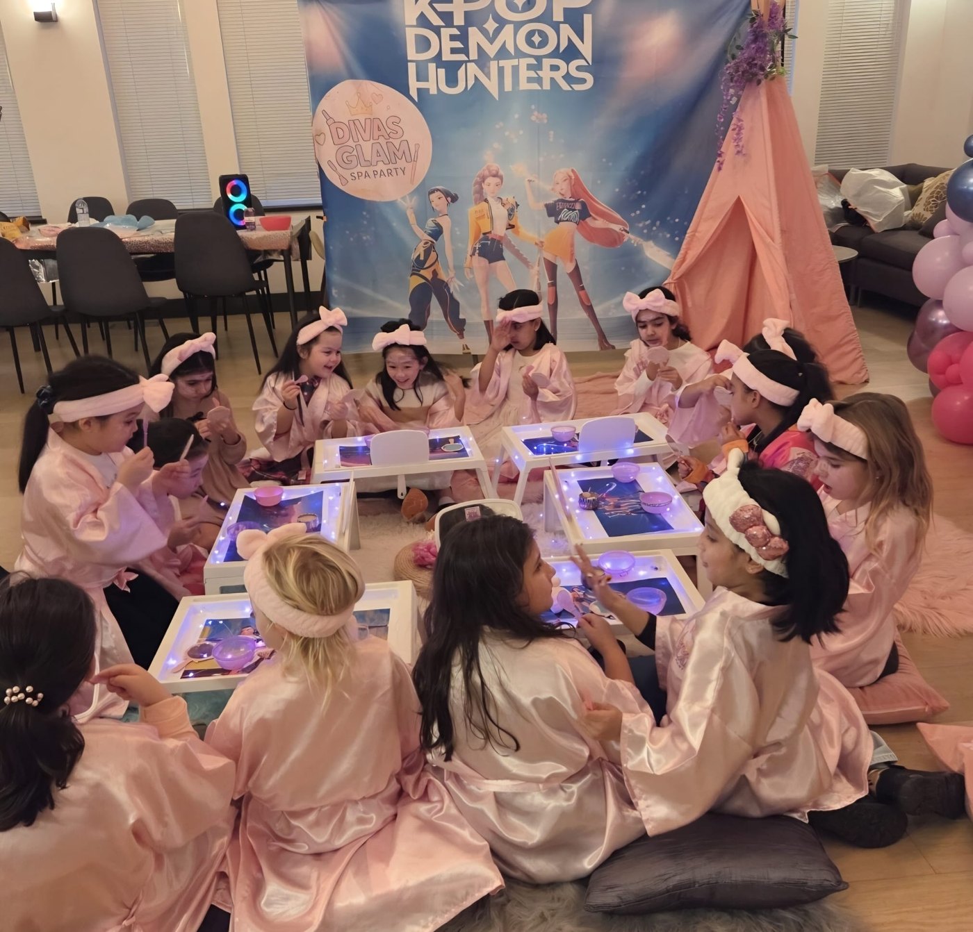 Group of girls in pink robes around white spa trays at a Divas Glam Spa Party with K-Pop themed backdrop